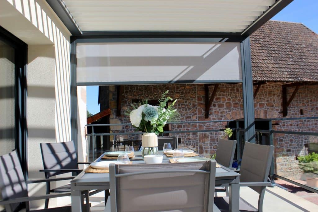 Pergola bioclimatique M130 avec store latéral blanc relevé sur une terrasse en pierre